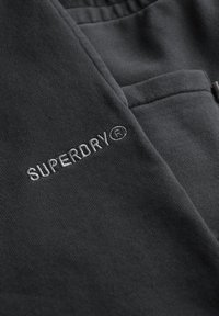 Donkergrijs sweatshirtmateriaal met gestikte "SUPERDRY"-logo. Zachte textuur met geribde tailleband en enkele zakdetails.