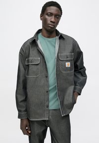 Koszula dżinsowa w ciemnoszarym kolorze z białym przeszyciem, dwoma przednimi kieszeniami i naszywką z logo. Noszona na turkusowej koszulce. Materiał bez zagnieceń.