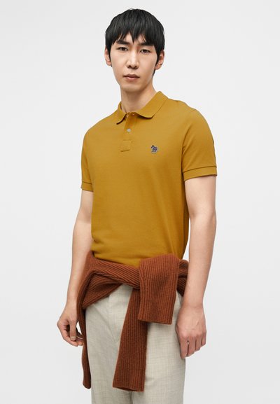 Μουσταρδί κίτρινο πόλο με δύο κουμπιά και ένα μικρό κεντημένο λογότυπο, συνδυασμένο με ανοιχτό μπεζ παντελόνι και σκουρόχρωμη καφέ μπλούζα δεμένη γύρω από τη μέση.