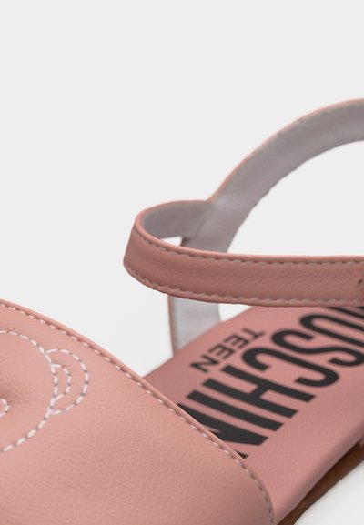 Ροζ σανδάλι με λουράκι και ορατό μαύρο τυπωμένο κείμενο στην εσωτερική σόλα, εμφανίζοντας λεπτομερείς ραφές στο πάνω μέρος.
