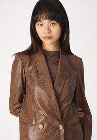 Kéttalpas barna műbőr blazer, kígyómintás textúrával, arany gombokkal és visszahajtott gallérral, áttetsző barna felsővel alatta.