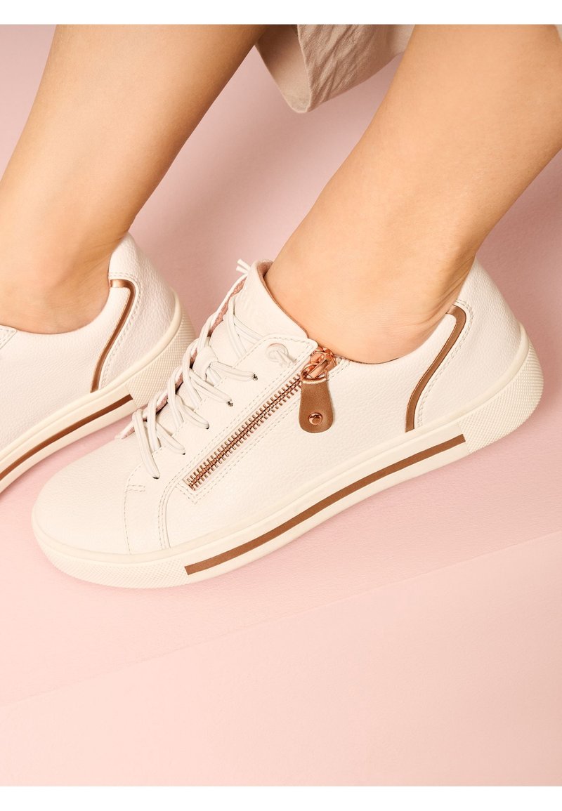 Pieds portant des baskets blanches décontractées avec fermeture éclair latérale et bandes accentuées marron sur des semelles en caoutchouc sur fond rose pâle.