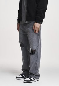 Osoba nosiaca voľné sivé roztrhané džínsy, čierno-biele tenisky, čiernu bundu a sivé tričko na bielom pozadí.