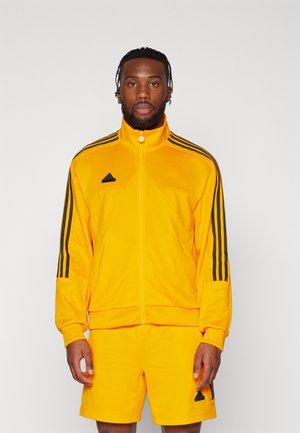 Tuta Adidas gialla con giacca con zip e pantaloncini, caratterizzata dal design a tre strisce nere e dettagli con il logo sul petto e sui pantaloncini.