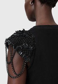 Czarny top bez rękawów z ozdobnym czarnym siatkowym detalem na ramionach, zdobionym aplikacjami w kształcie liści oraz zwisającymi czarnymi łańcuszkami.