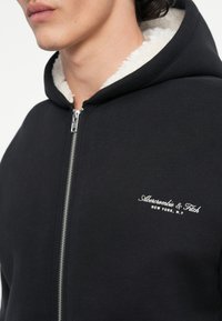 Hoodie noir zippé avec une doublure en sherpa blanche et un logo sur la poitrine. Dispose d'une fermeture éclair argentée et d'une capuche spacieuse. Tissu doux et texturé.