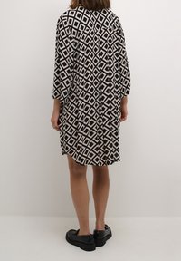 Sort og cremefarvet kjole med diamantmønster, løs pasform, trekvartærmer og åben ryg; kombineret med sorte sko.