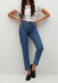 Jeans de mezclilla azul de cintura alta con corte recto, que cuentan con un cierre de botón y cremallera, y un desgaste mínimo. Combinados con mocasines negros.