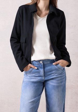 Mujer con chaqueta negra con cremallera sobre una camiseta blanca, con las manos en los bolsillos de unos vaqueros azul claro, frente a un fondo neutro.