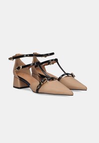 Zapatos de punta beige con tiras negras brillantes y acentos dorados. Cuentan con un tacón bajo y resistente y un diseño de barra T para mayor soporte.