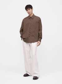 Homme debout portant une chemise à rayures verticales marron et beige, un pantalon blanc ample et des chaussures marron foncé sur un fond blanc uni.