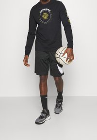 Czarny longsleeve z grafiką "Warriors" i żółtymi akcentami, zestawiony z czarnymi szortami z białym logo Nike, trzymający piłkę koszykową.