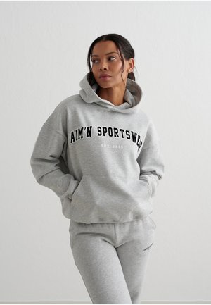 Donna con capelli scuri che indossa una felpa con cappuccio e pantaloni della tuta grigio chiaro con la scritta "AIM'N SPORTSWEAR", in piedi davanti a uno sfondo bianco semplice.