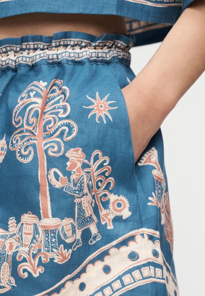 Χέρι με μπλε ένδυμα στην τσέπη, διακοσμημένο με λευκά και ροδακινί παραδοσιακά λαϊκά μοτίβα δέντρων, ήλιου και ανθρώπινων φιγούρων που παίζουν μουσικά όργανα.