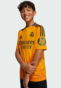 Maillot de soccer orange avec des accents noirs, arborant les logos "Emirates" et "hp". Texture lisse avec des rayures diagonales et un col rond.