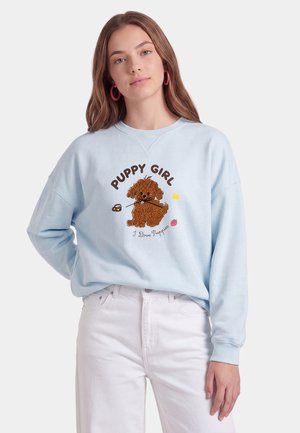 Giovane donna con lunghi capelli castani indossa una felpa azzurro chiaro con grafica di cucciolo marrone e testo «"Puppy Girl"», abbinata a pantaloni bianchi.