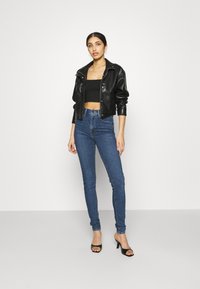Fekete műbőr rövid dzseki álló gallérral, fekete crop top-pal és kék skinny farmernadrággal, valamint fekete nyitott orrú magassarkú cipővel párosítva.