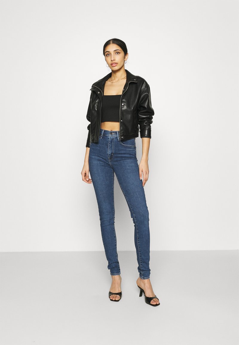 Chaqueta cropped de piel sintética negra con cuello de solapa, combinada con un top corto negro y jeans skinny azul, y tacones abiertos negros.