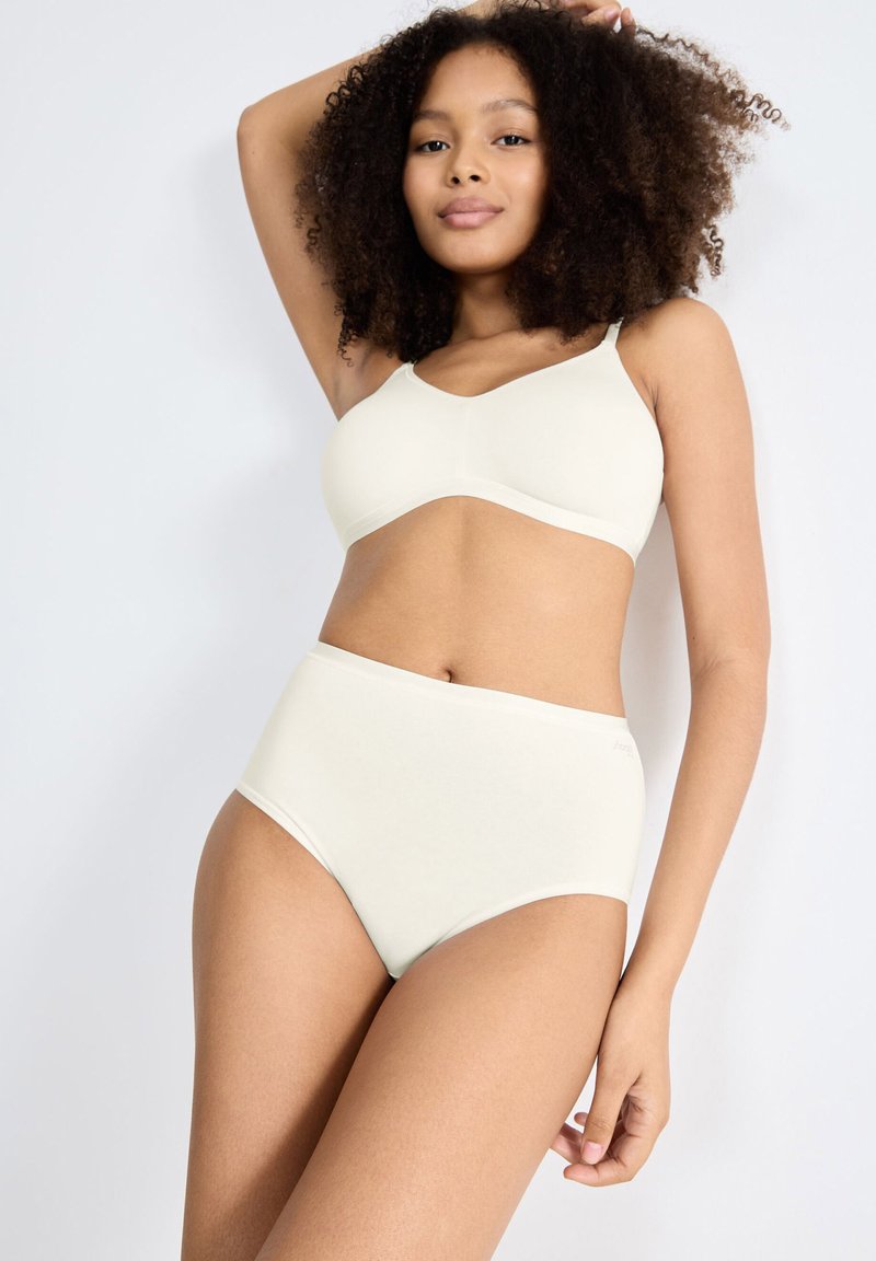 Vrouw met krullend haar poseert in een naadloze beige high-waisted ondergoedset tegen een effen lichte achtergrond.