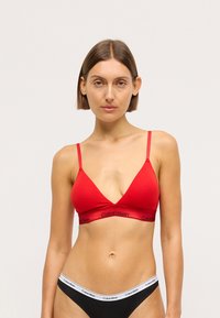 Raudona medvilninė trikampė liemenėlė su plonais diržais ir elastinga juosta, su juodu "Calvin Klein" logotipu. Atitinkamos juodos maždaug juosmeninės bikinio kelnaitės su logotipo juosta.