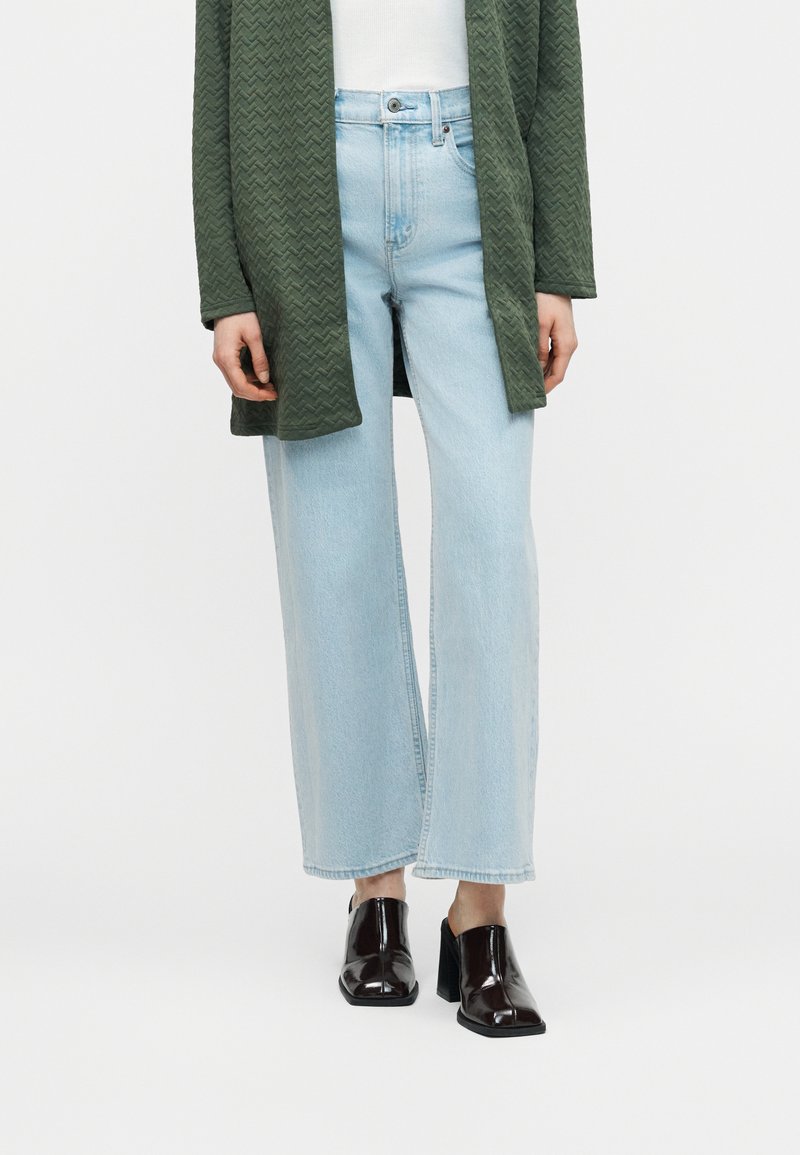 Φαρδιά τζιν παντελόνια σε ανοιχτό μπλε χρώμα, συνδυασμένα με μαύρα mules με τακούνι και μακρύ πλεκτό πράσινο σκούρο ζακέτα πάνω από λευκό τοπ.