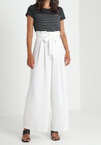 Kobieta w czarno-białym pasiaku, w spodniach z wysokim stanem i szerokimi nogawkami w kolorze białym, z przewiązanym paskiem oraz czarnych sandałach na obcasie z odkrytymi palcami.