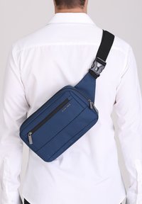 Borsa crossbody blu navy realizzata in tessuto testurizzato. Presenta due scomparti con zip e una tracolla nera regolabile. Design compatto e geometrico.