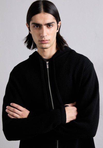 Schwarzer Kapuzenpullover mit Reißverschluss vorne, der eine weiche Textur und ein lässiges Design aufweist. Das Modell hat die Arme verschränkt und zeigt die Passform des Kleidungsstücks.