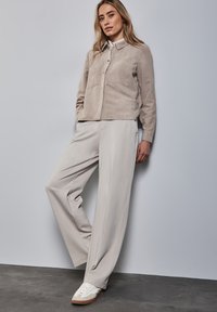Beige suède knoopshirt met twee borstzakken, gecombineerd met lichtgrijze wijde broek en witte sneakers met bruine accenten.