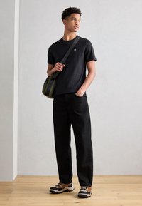 Crna pamučna majica s malim logom, uparen s opuštenim crnim hlačama. Dodaci uključuju maslinasto zeleni torbu za rame i pošivene cipele.