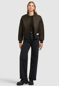 Olive zelená bomber bunda s rebrovanými manžetami a lemom, so zipsom a bočnými vreckami, skombinovaná s čiernymi džínsami so zvýšeným pásom a bootami.