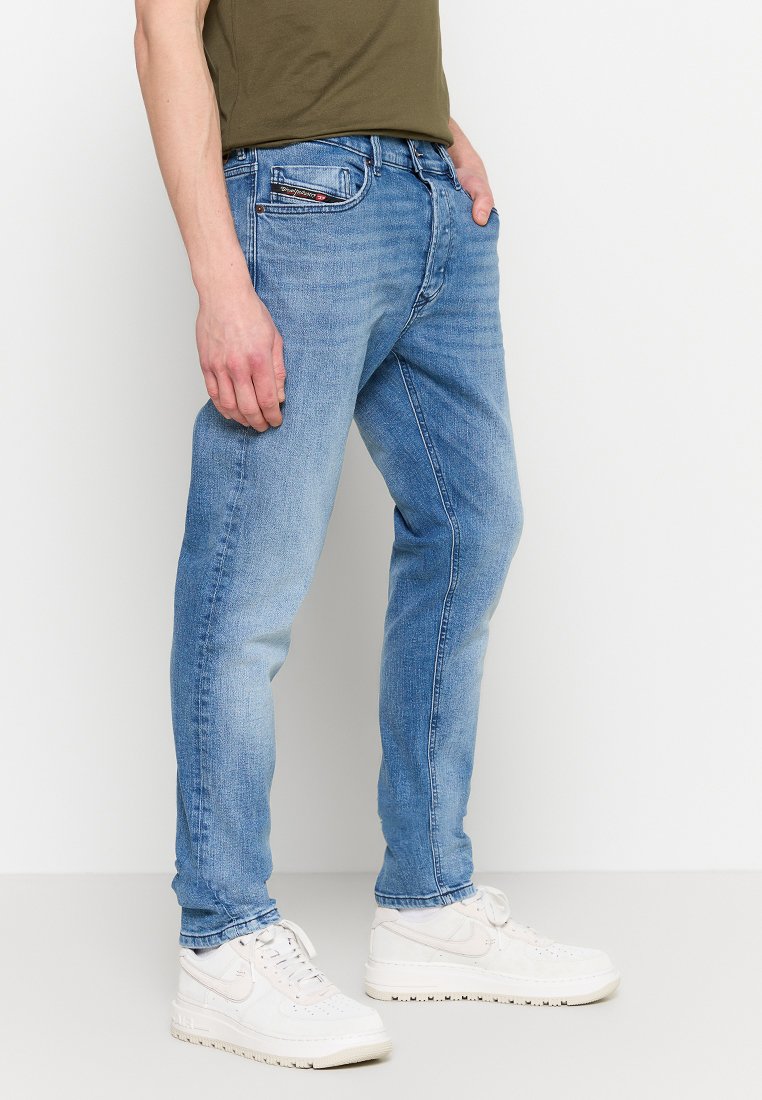 Homme portant un jean bleu clair et des baskets blanches, se tenant contre un fond blanc uni.