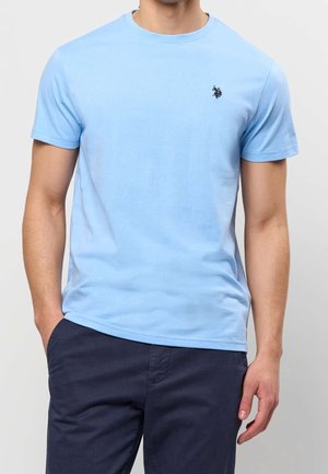 Homme portant un t-shirt bleu clair à manches courtes avec un petit logo foncé sur la poitrine et un pantalon bleu marine foncé, une main dans la poche.