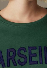 Nahaufnahme des Nackens und des oberen Brustbereichs, der ein grünes Shirt mit großen, dunkelblauen gestickten Buchstaben trägt, die "ARSEI" buchstabieren.