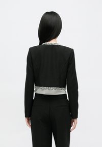 Μαύρο σακάκι μ ε δομημένο σχεδιασμό, που διαθέτει ασημί διακοσμημένο γιακά και αντίθετη ζώνη σατέν στη μέση. Η πίσω όψη αναδεικνύει τη ραμμένη εφαρμογή.