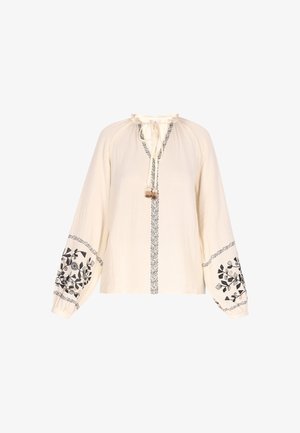 Blusa color crema con mangas largas y recogidas. Presenta bordados florales negros en las mangas y un escote con lazo y borlas. Tejido texturizado.