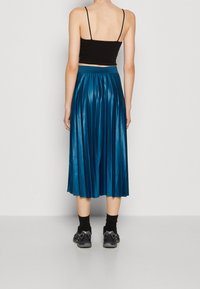 Gonna midi plissettata turchese con finitura lucida, abbinata a un top nero corto e sneaker nere, vista da dietro.