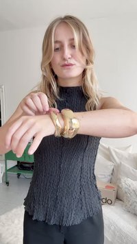 Kaksi paksua kultaketjua, joissa on sileä, heijastava pinta, jotka on puettu ranteeseen. Yläosan ketjussa on virtaava, aaltoileva muotoilu.