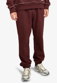 Bordeaux sweatpants met elastische boorden, gemaakt van een zachte stof. Opvallend witte stiksels. Gecombineerd met grijze en witte sportschoenen.