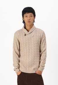 Pull en maille beige avec un motif de cable, col châle et un détail de bouton unique. Présente des poignets et un ourlet côtelés. Porté sur un pantalon marron.