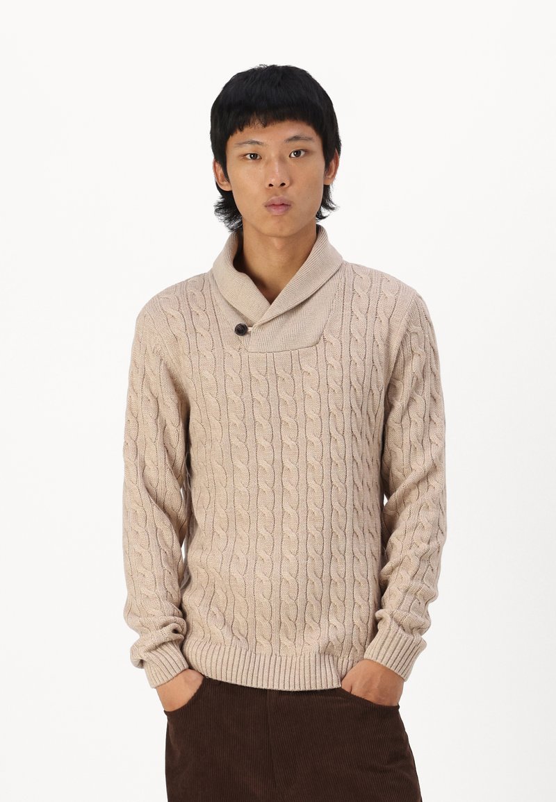 Pull en maille beige avec un motif de cable, col châle et un détail de bouton unique. Présente des poignets et un ourlet côtelés. Porté sur un pantalon marron.