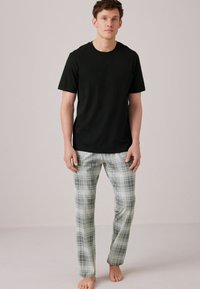 Black cotton t-shirt paired with light green plaid pajama pants featuring black and grey lines; model stands barefoot against a plain background.