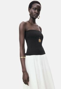 Schwarzes, figurbetontes, trägerloses Oberteil, kombiniert mit einem weißen, fließenden Rock. Goldene Accessoires betonen den Look und schaffen einen auffälligen Kontrast.