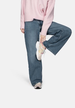 Person trägt weit geschnittene blaue Jeans, pinkfarbenen Pullover und beige Turnschuhe, hebt ein Bein an und hält den Schuh mit der Hand fest.