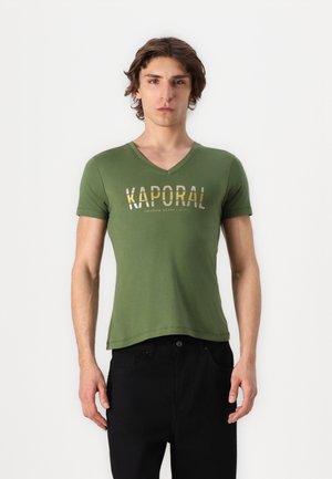 Jonge man staand, gekleed in een groen V-hals T-shirt met de tekst "KAPORAL" en een zwarte broek, tegen een effen witte achtergrond.