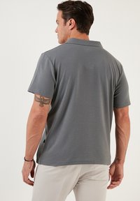 Polo gris de manga corta, con tejido texturizado y cuello plano. Presenta una abertura lateral y una sutil etiqueta de la marca en el dobladillo. Se muestra vista trasera.