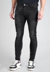 Jean en denim noir à coupe slim, présentant des détails usés et une texture lisse, associés à des baskets noires et blanches.