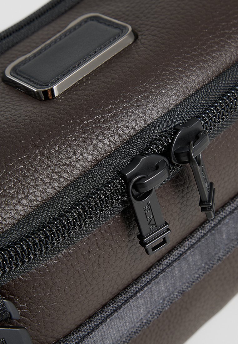 Die schwarze Ledertasche verfügt über ein strukturiertes Material, doppelte schwarze Reißverschlüsse und einen grauen Akzentgurt. Sie enthält ein rechteckiges Logopatch oben.