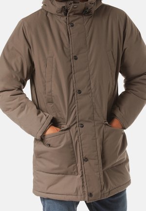 Mężczyzna w brązowej pikowanej zimowej kurtce z kapturem i rękami w przednich kieszeniach, widoczne niebieskie dżinsy.