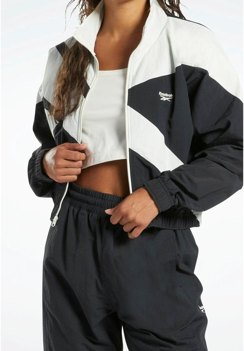 Svart och vit Reebok-jacka med geometrisk design, dragkedjestängning och elastiska ärmslut, i kombination med svarta byxor med dragsko. Slät tygtextur.